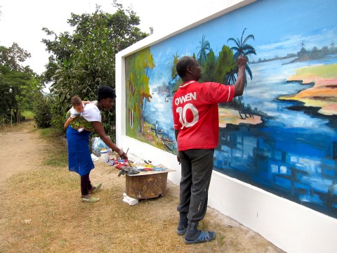 20 août 2012 › Portant son fils Quentin sur le dos, Rhode Makoumbou réalise la fresque murale «Le village de pêcheurs».